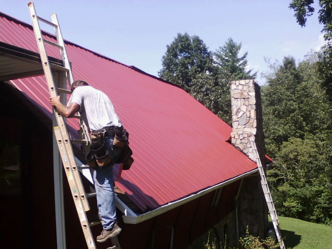 Roofer on a ladder beside a red metal roof and stone chimney — Soffit & Fascia Repair by Roofing Brothers in Cape Charles